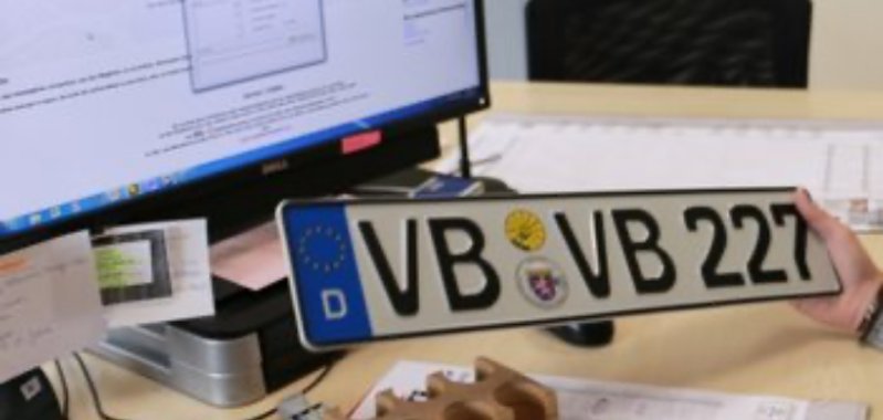 Ein Autokennzeichen wird in die Kamera gehalten. Im Hintergrund ist ein Büroarbeitsplatz zu sehen