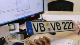 Symbolbild Zulassungsbehörde Ein Autokennzeichen wird in die Kamera gehalten. Im Hintergrund ist ein Büroarbeitsplatz zu sehen