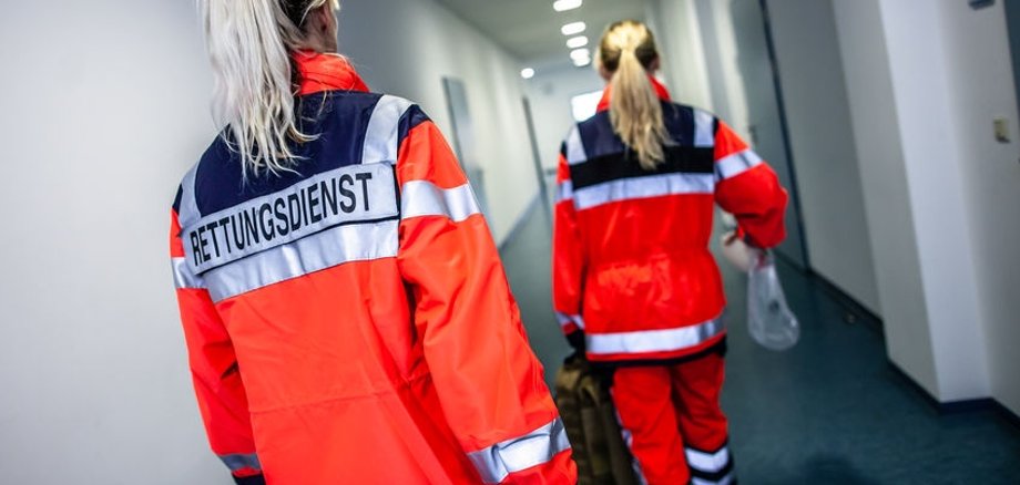 Rettungsdienstmitarbeiterinnen in einem langen Gang auf dem Weg zum Einsatz. Sie sind von hinten zu sehen und tragen auffällig rote Jacken. 