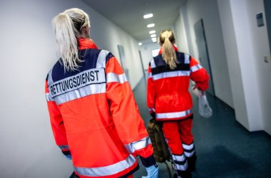 Rettungsdienstmitarbeiterinnen in einem langen Gang auf dem Weg zum Einsatz. Sie sind von hinten zu sehen und tragen auffällig rote Jacken. 