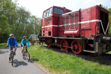 Zwei Radfahrer fahren auf einem Radweg. Sie haben blaue Jacken an und fahren an einer stillgelegten Lok vorbei. 