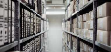 Blick in eine Regalreihe in einem Archiv. Regale sind vollgestellt mit Kisten und Ordnern. 