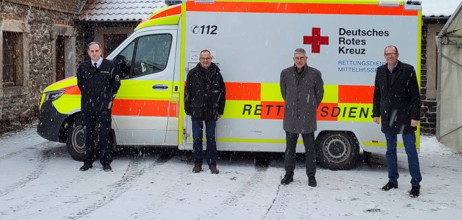 Kreisbrandinspektor (von links) Dr. Sven Holland, RDMH-Geschäftsführer Markus Müller, Landrat Manfred Görig und Bürgermeister Sascha Spielberger beim Ortstermin an der Rettungswache in Freiensteinau. Sie stehen vor einem Rettungswagen. Es schneit, der WAgen steht vor einem Bruchsteingebäude