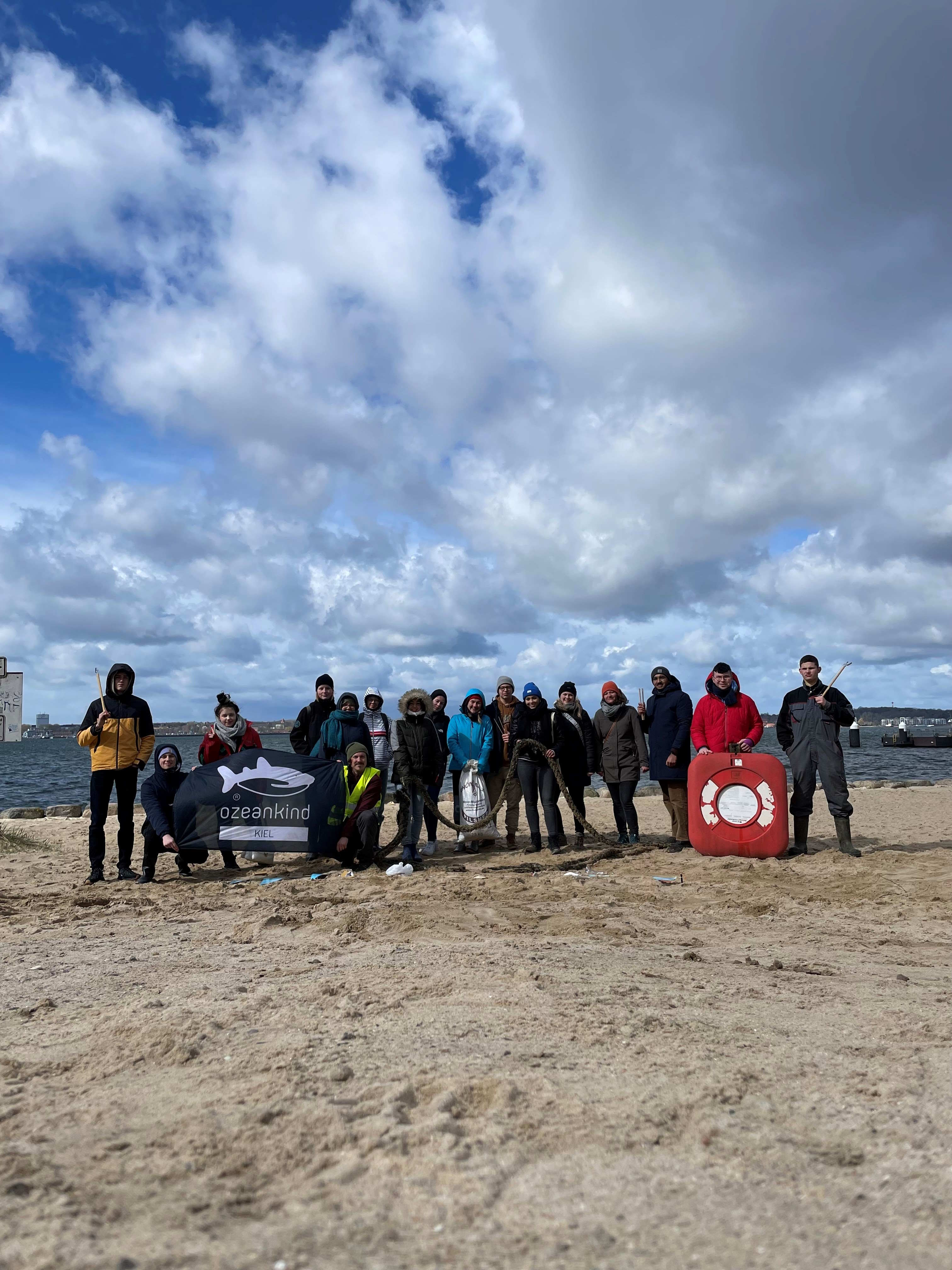 Am Strand bei Kiel sammeln die Mitglieder des Kreisjugendparlaments in nur einer Stunde 32 Kilogramm Müll ein. Auf dem Foto ist ein Strand, eine Menschengruppe und im Hintergrund das Meer zu sehen. 