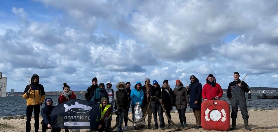 Am Strand bei Kiel sammeln die Mitglieder des Kreisjugendparlaments in nur einer Stunde 32 Kilogramm Müll ein. Auf dem Foto ist ein Strand, eine Menschengruppe und im Hintergrund das Meer zu sehen. 