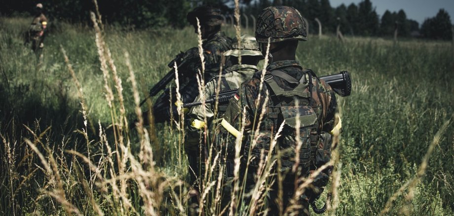 Soldaten in Tarnkleidung laufen durchs hohe Gras. Sie laufen mit dem Rücken zur Kamera.