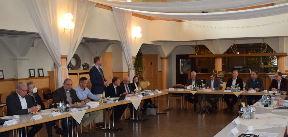 in einem Festsaal sitzen in U-förmiger Runde Menschen in einer Besprechung. Der Erste Kreisbeigeordnete Dr. Jens Mischak (stehend) begrüßte die Gäste aus Wiesbaden sowie Nord- und Mittelhessen in der Kreisstadt. Kreisstraßen und Mobilität in ländlich strukturierten Kreisen stand im Mittelpunkt. 