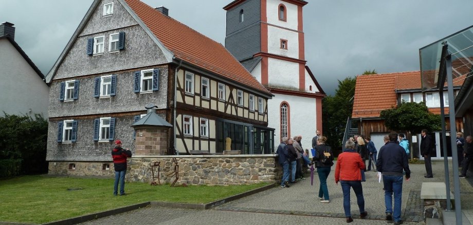 Eine Impression der Bereisung im Jahr 2017. Die Kommission machte bei ihrer Bereisung in Hopfmannsfeld Halt. vor einem Fachwerkhaus hat sich eine Gruppe Menschen versammelt. Im Hintergrund ist ein weiß-roter Kirchturm zu sehen.