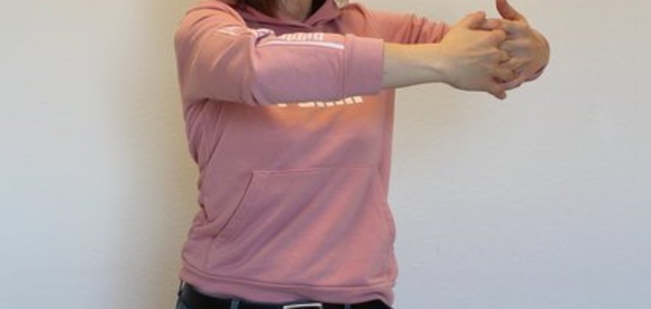 Eine Frau in pinkem Sweatshirt macht eine Übung für die Wirbelsäule vor. 
