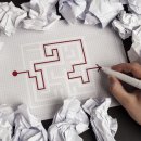 Auf einem karierten Zettel malt eine Hand mit einem roten Stift eine Strecke durch ein Labyrinth nach. Rund um das Blatt liegen Papierknäuel