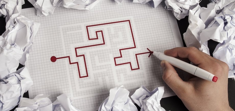Auf einem karierten Zettel malt eine Hand mit einem roten Stift eine Strecke durch ein Labyrinth nach. Rund um das Blatt liegen Papierknäuel