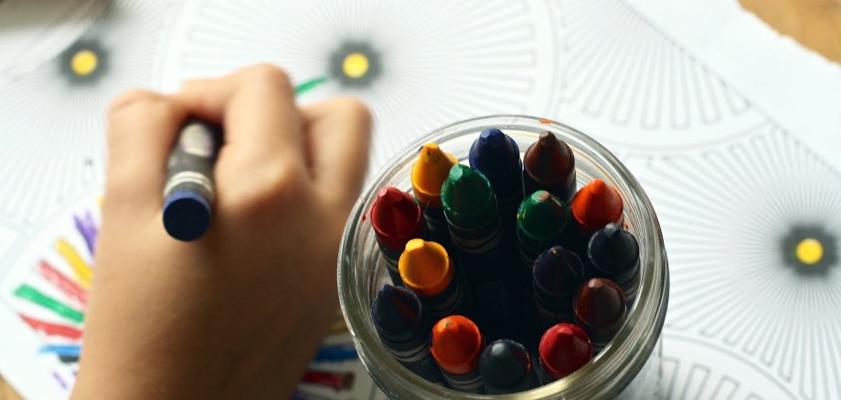Eine Kinderhand malt mit Wachsmalstiften Mandalas aus. Von oben fotografiert ist auch ein Glas mit verschiedenen Stiften zu sehen