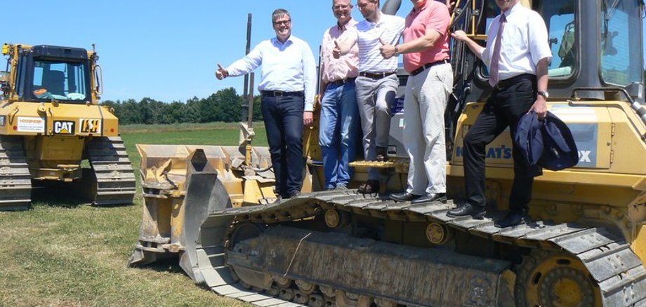 Fünf Männer stehen auf einer KEttenraupe, die eine Baustelle vorbereiten wird. Daumen hoch: Mückes ehemaliger Bürgermeister Matthias Weitzel, Thomas Heidlas, der Fachbereichsleiter Bauen und Liegenschaften der Gemeinde, Erster Kreisbeigeordneter Dr. Jens Mischak, Landrat Manfred Görig und Mückes Bürgermeister Andreas Sommer freuen sich, dass die Bauarbeiten am Montag starten.