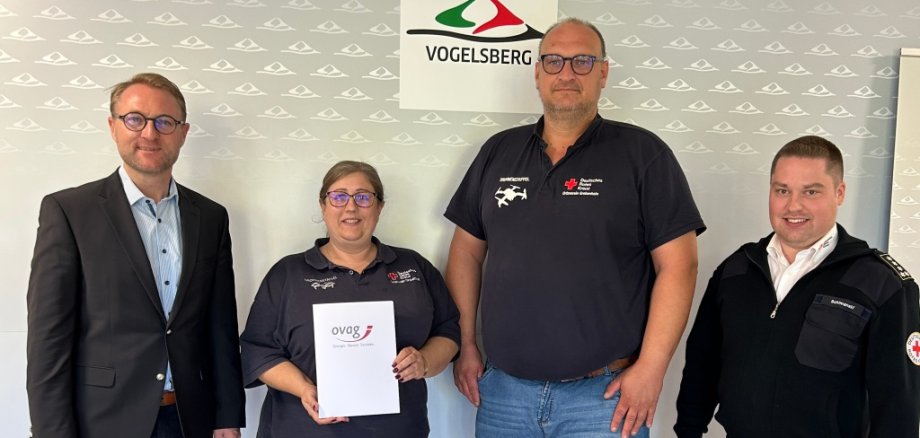 Vier Personen stehen vor einer Fotowand und präsentieren einen Spendenbescheid. Sie lächeln in die Kamera.
