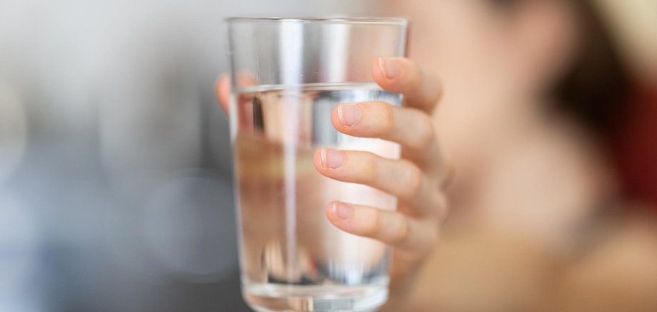 Eine Frau hält ein Glas Wasser in die Kamera. Der Hintergrund ist verschwommen