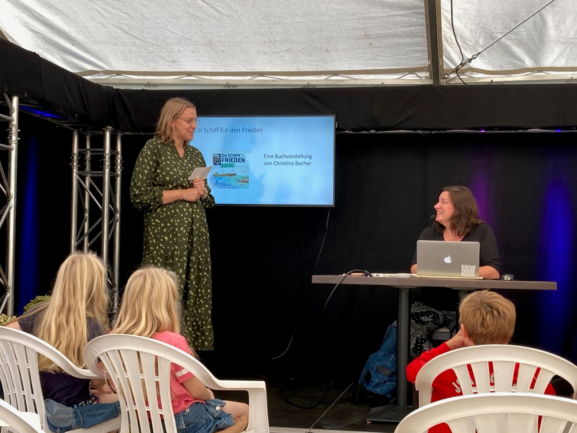 Zwei Frauen stehen auf einer kleinen Bühne und sprechen miteinander. Im Publikum sind Kinder zu sehen, die beim Erzählfestival in Homberg lauschen.