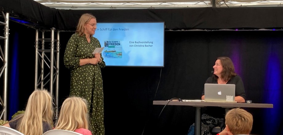 Zwei Frauen stehen auf einer kleinen Bühne und sprechen miteinander. Im Publikum sind Kinder zu sehen, die beim Erzählfestival in Homberg lauschen.