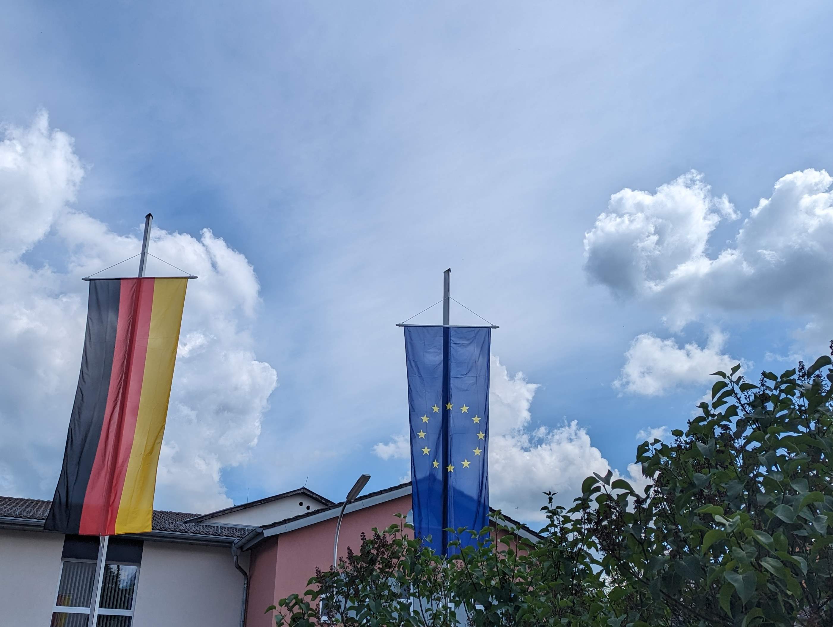 EU- und Deutschland-Flaggen an zwei Fahnenmasten. Dahinter sind blauer Himmel und Wolken zu sehen.