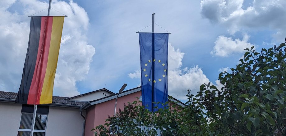 EU- und Deutschland-Flaggen an zwei Fahnenmasten. Dahinter sind blauer Himmel und Wolken zu sehen.
