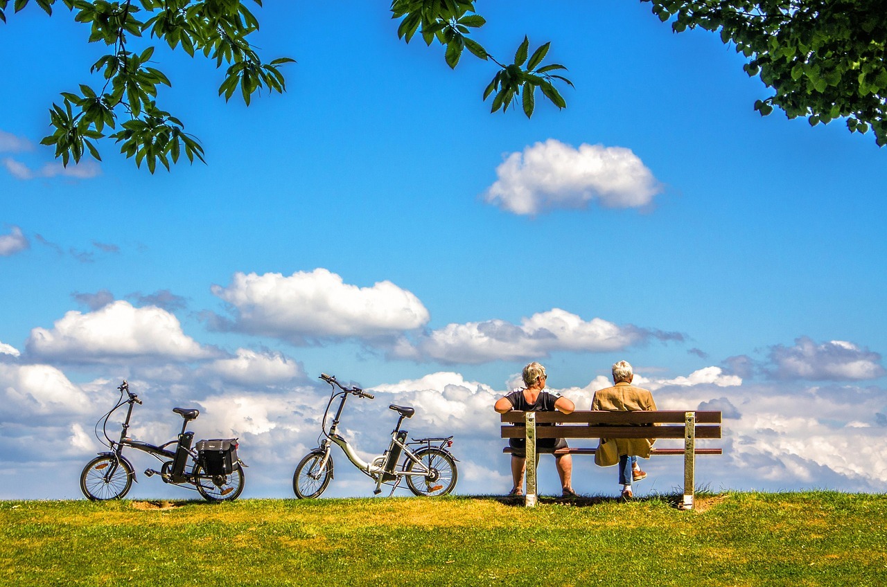 ein Seniorenehepaar sitzt auf einer Bank und blickt auf blauen Himmel. Links sind zwei E-Bikes zu sehen.