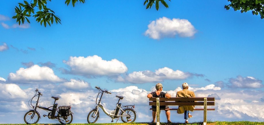 ein Seniorenehepaar sitzt auf einer Bank und blickt auf blauen Himmel. Links sind zwei E-Bikes zu sehen.