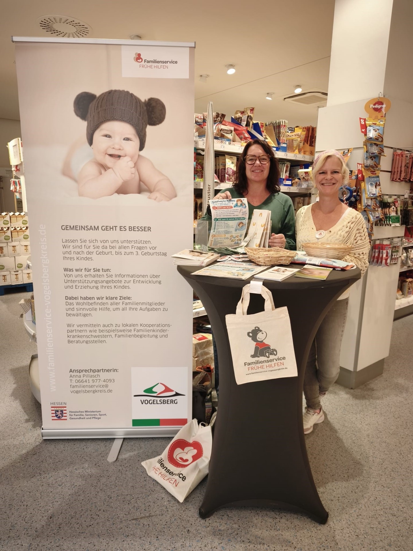 Zwei Frauen stehen an einem Stehtisch in einer Drogerie und informieren zu Hilfsangeboten für junge Eltern.