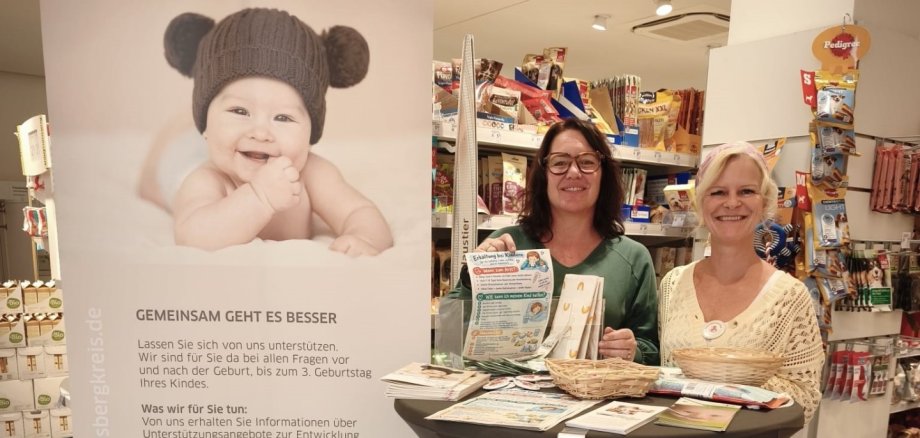 Zwei Frauen stehen an einem Stehtisch in einer Drogerie und informieren zu Hilfsangeboten für junge Eltern.