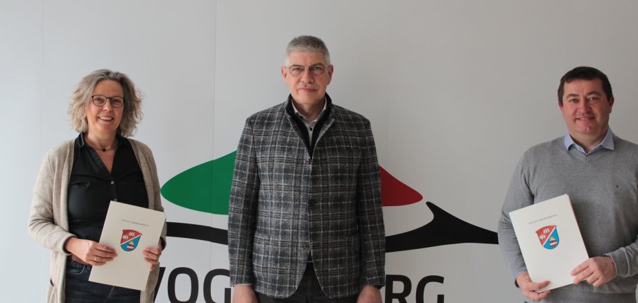 Landrat Manfred Görig gratuliert Ulrike Geisel und Bernd Ruhl zum 25-jährigen Dienstjubiläum. Sie stehen vor einer weißen Fotowand