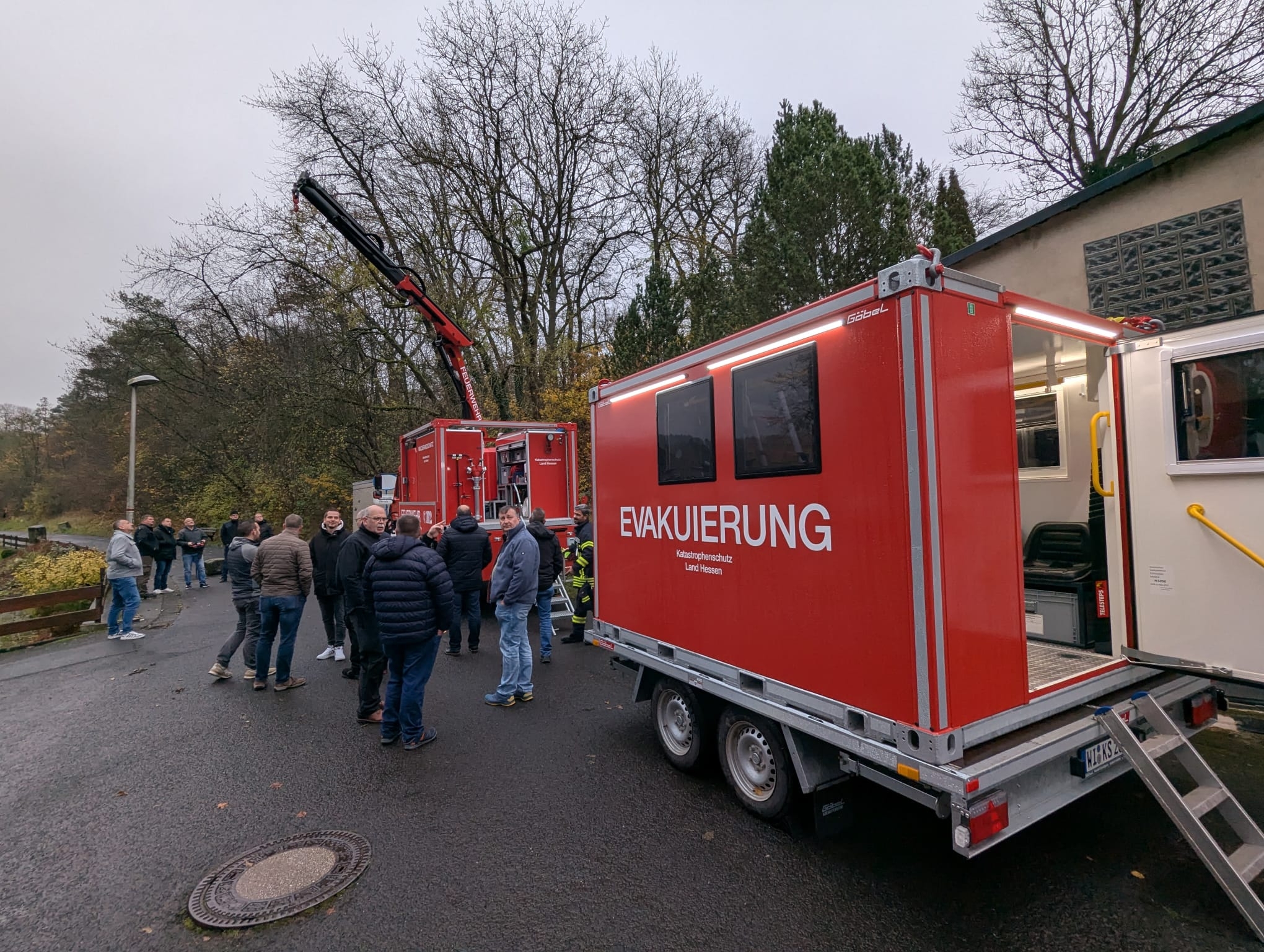 Mehrere Personen inspizieren Spezialgerät der Feuerwehr. Es ist auch ein Zugfahrzeug mit Kranausleger zu sehen. 