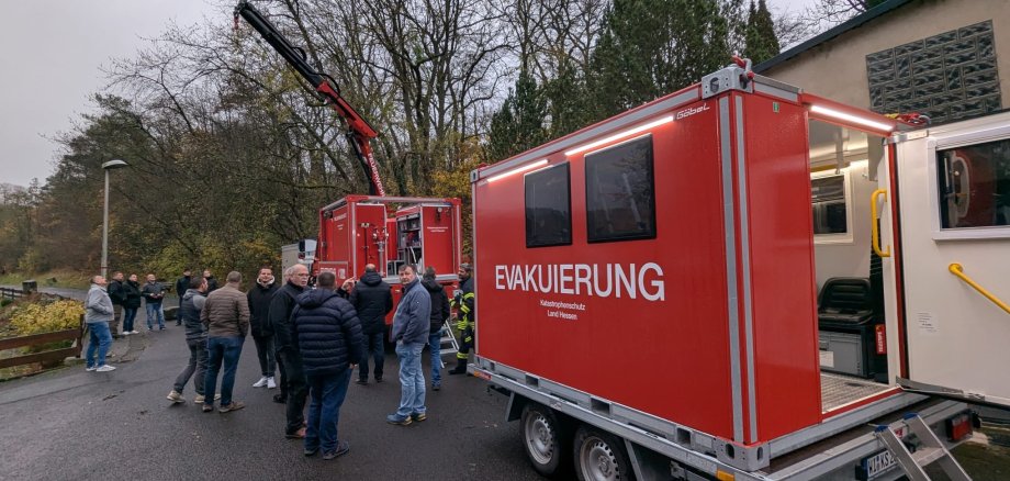 Mehrere Personen inspizieren Spezialgerät der Feuerwehr. Es ist auch ein Zugfahrzeug mit Kranausleger zu sehen. 