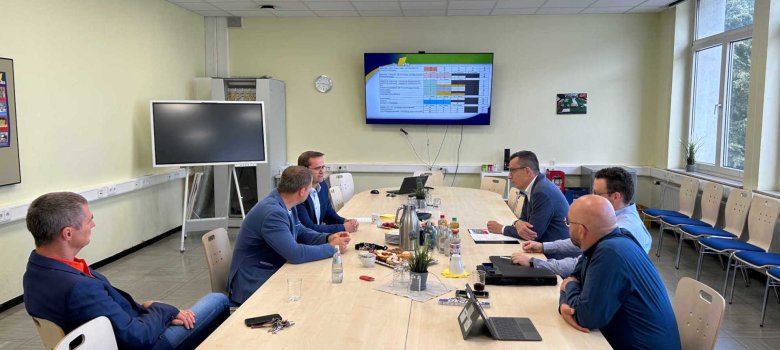 Mehrere Personen sitzen an einem Konferenztisch und sprechen miteinander. Im Hintergrund ist ein großer Bildschirm zu sehen.