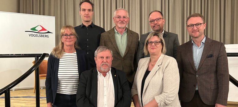 Sieben Personen haben sich auf einer Treppe in einem Saal zum Gruppenbild aufgestellt.