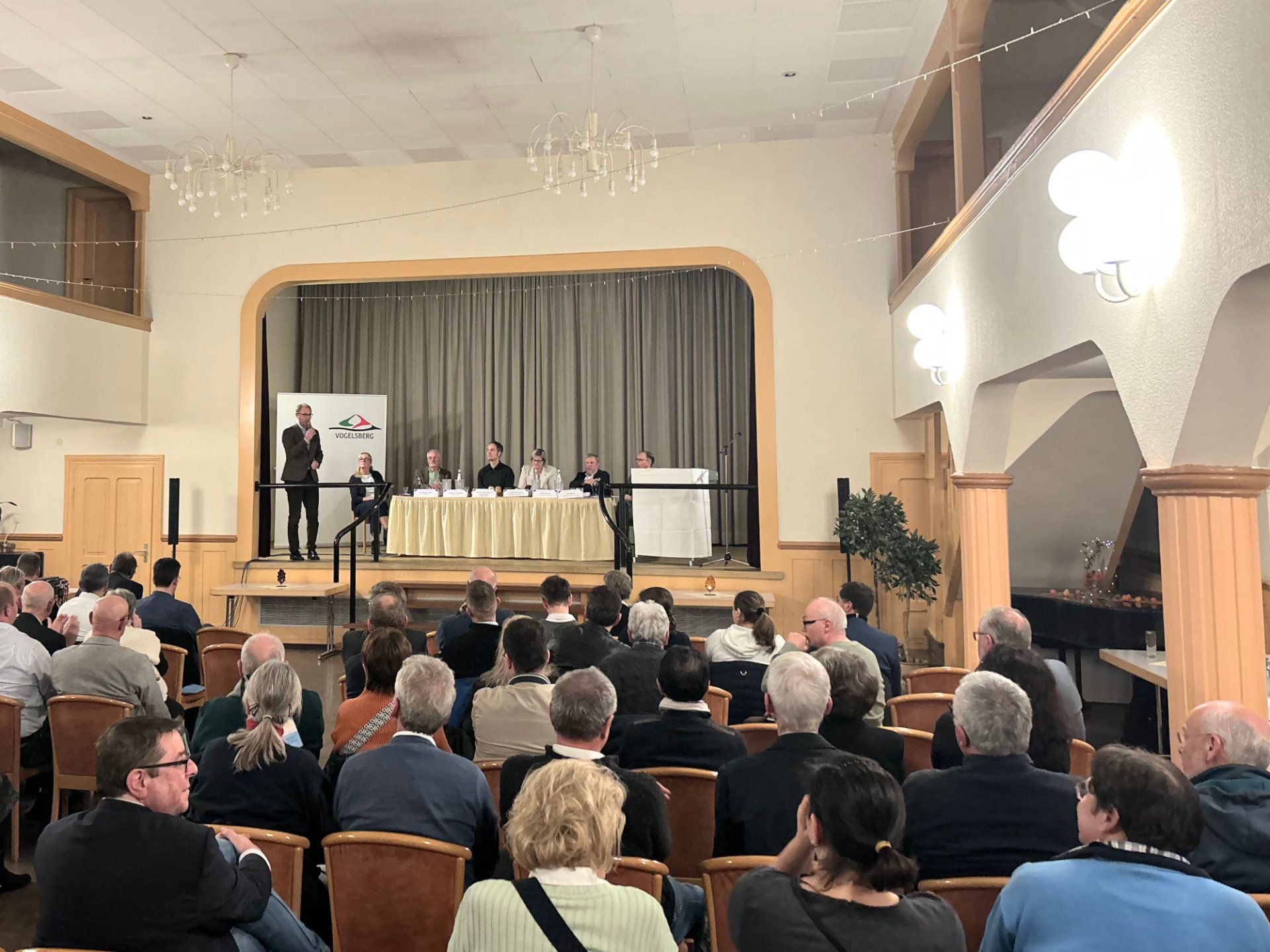 In einem Saal sind rund 90 Menschen zu sehen, die in Reihen sitzen. Auf einer Bühne diskutieren mehrere PErsonen. 