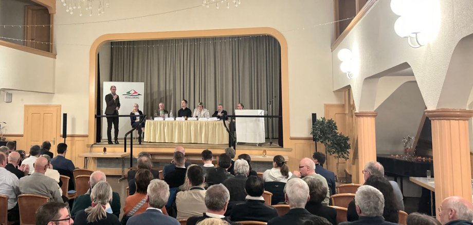 In einem Saal sind rund 90 Menschen zu sehen, die in Reihen sitzen. Auf einer Bühne diskutieren mehrere PErsonen. 