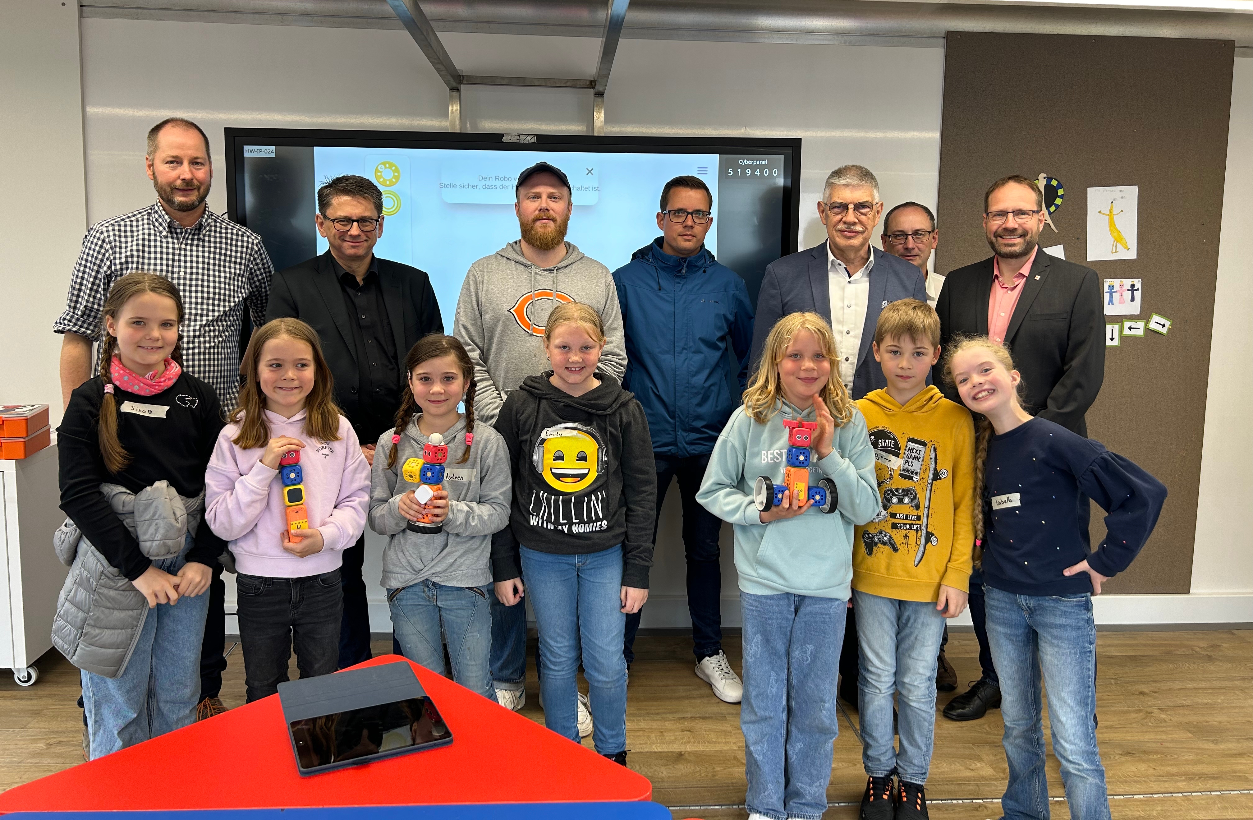 Erwachsene und Kinder stehen vor einer digitalen Tafel. Die Kinder halten selbst gestaltete Roboter in den Händen.