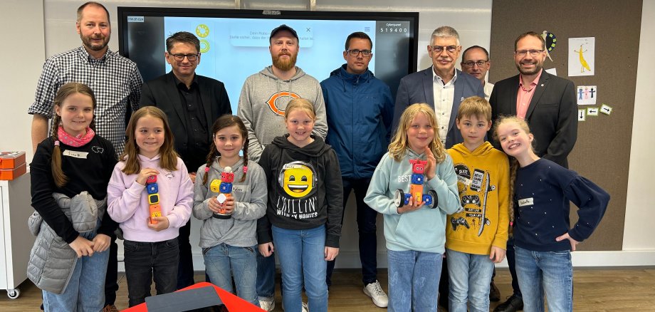 Erwachsene und Kinder stehen vor einer digitalen Tafel. Die Kinder halten selbst gestaltete Roboter in den Händen.