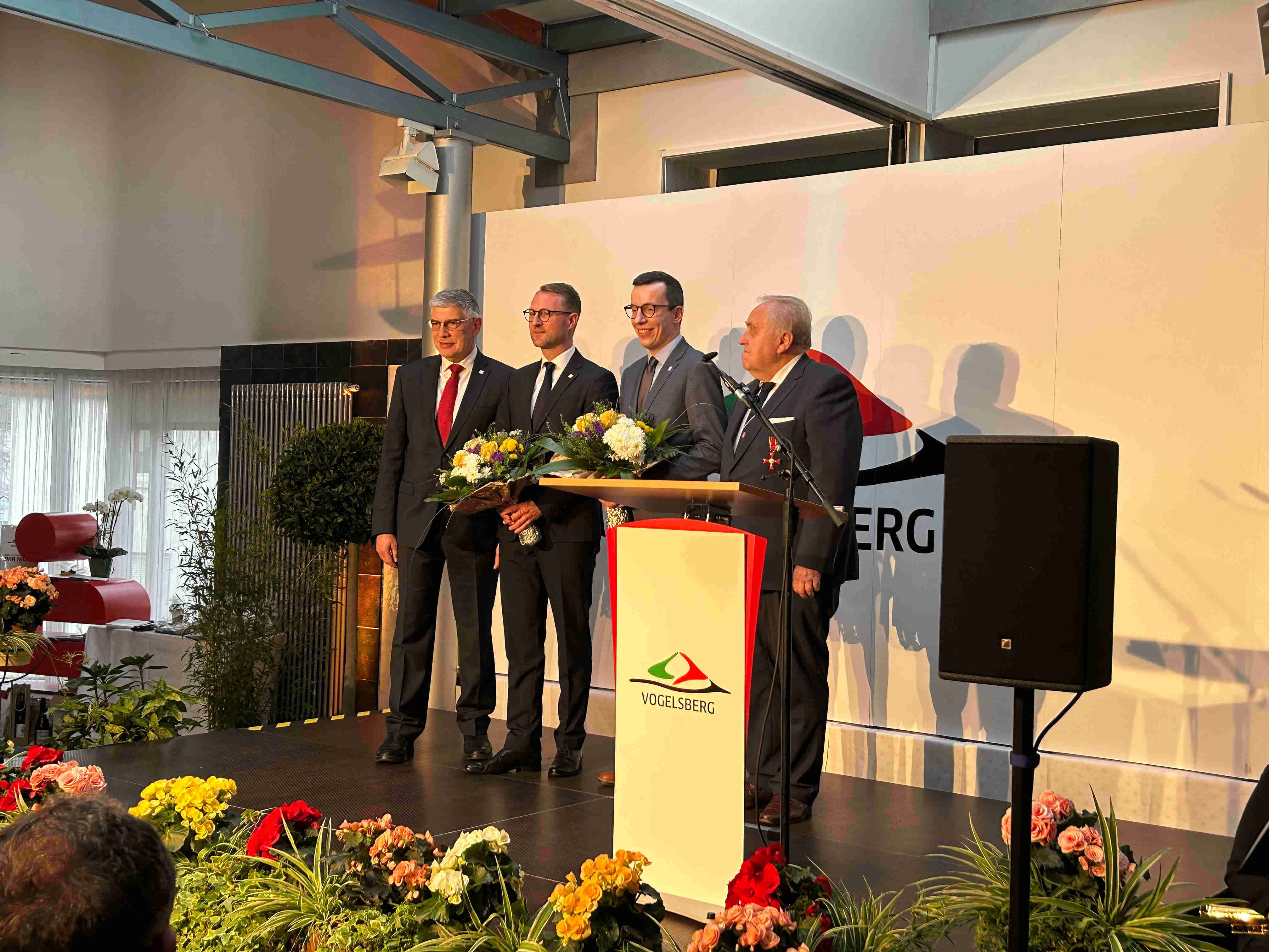 Manfred Görig, Dr. Jens Mischak, Patrick Krug und Dr. Hans Heuser stehen auf einer Bühne vor einer weißen Wand. Sie tragen Anzüge und lächeln in die Kamera