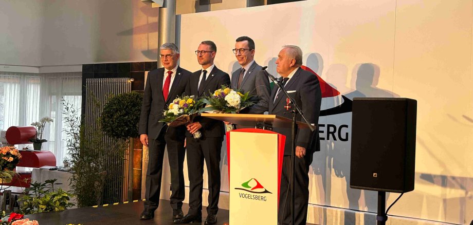 Manfred Görig, Dr. Jens Mischak, Patrick Krug und Dr. Hans Heuser stehen auf einer Bühne vor einer weißen Wand. Sie tragen Anzüge und lächeln in die Kamera