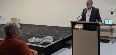 Landrat Manfred Görig steht an einem hell lackierten Rednerpult. Links von ihm ist eine kleine Bühne zu sehen. Er spricht zu Menschen in einer Schulaula