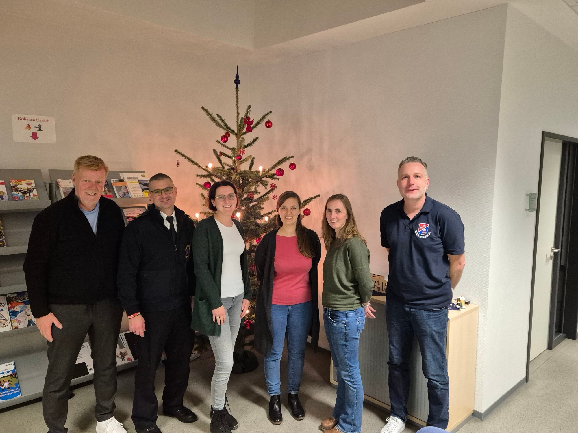 sechs Menschen haben sich vor einem geschmückten Weihnachtsbaum zum Gruppenbild aufgestellt und lächeln in die Kamera