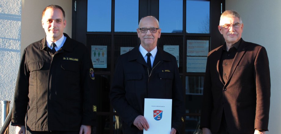 Drei Herren stehen vor einer Eingangstür auf einer Treppe. Der Herr in der Mitte hält eine Urkunde in seinen Händen. Dieses Jubiläum hat Seltenheitswert: Ein Vierteljahrhundert war der Homberger Peter Pfeil (Mitte) als Kreisbrandmeister tätig, jetzt wurde er von Landrat Manfred Görig und Kreisbrandmeister Dr. Sven Holland verabschiedet. 