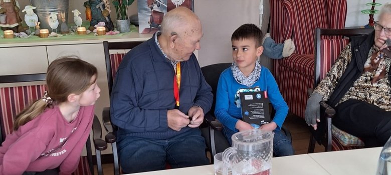 Ein Senior und zwei Kinder sitzen an einem Tisch und sind in ein Gespräch vertieft. Ein Junge hat gerade aus einem Buch vorgelesen.