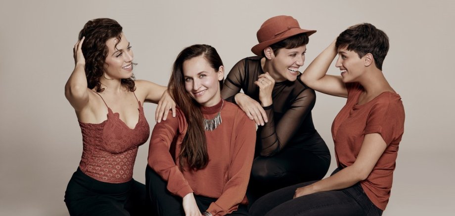 Vier Frauen sitzen gemeinsam auf dem Boden und lächeln in die Kamera. 
