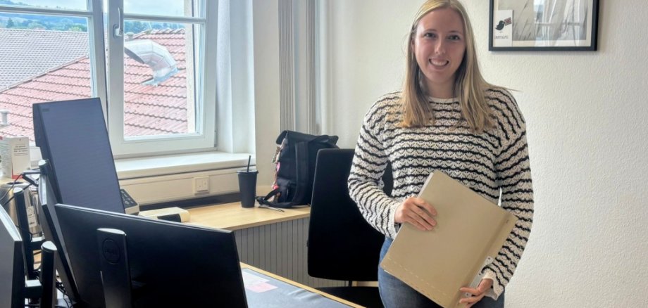 Luisa Schmadel arbeitet als gelernte Justizfachangestellte seit 2023 beim Vogelsbergkreis im Bereich „Hilfe zur Pflege“ im Amt für Soziales und Ausländerrecht. Sie steht hinter ihrem Schreibtisch, hat einen Ordner in der Hand und lächelt in die Kamera.