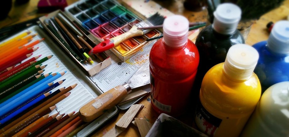 Viele Mal-Utensilien sind auf einem Tisch verteilt. Es stehen Farbflaschen, Stifte und Pinsel verteilt auf dem Tisch
