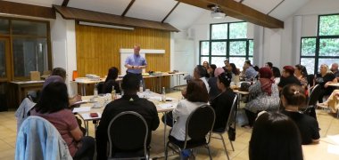 Björn Preuß-von Brincken, Kreisbrandmeister und Interkultureller Berater Feuerwehr im Austausch mit den Ehrenamtlichen im Sprachmittlungspool. Sie sitzen in einem Saal an einigen Tischen.