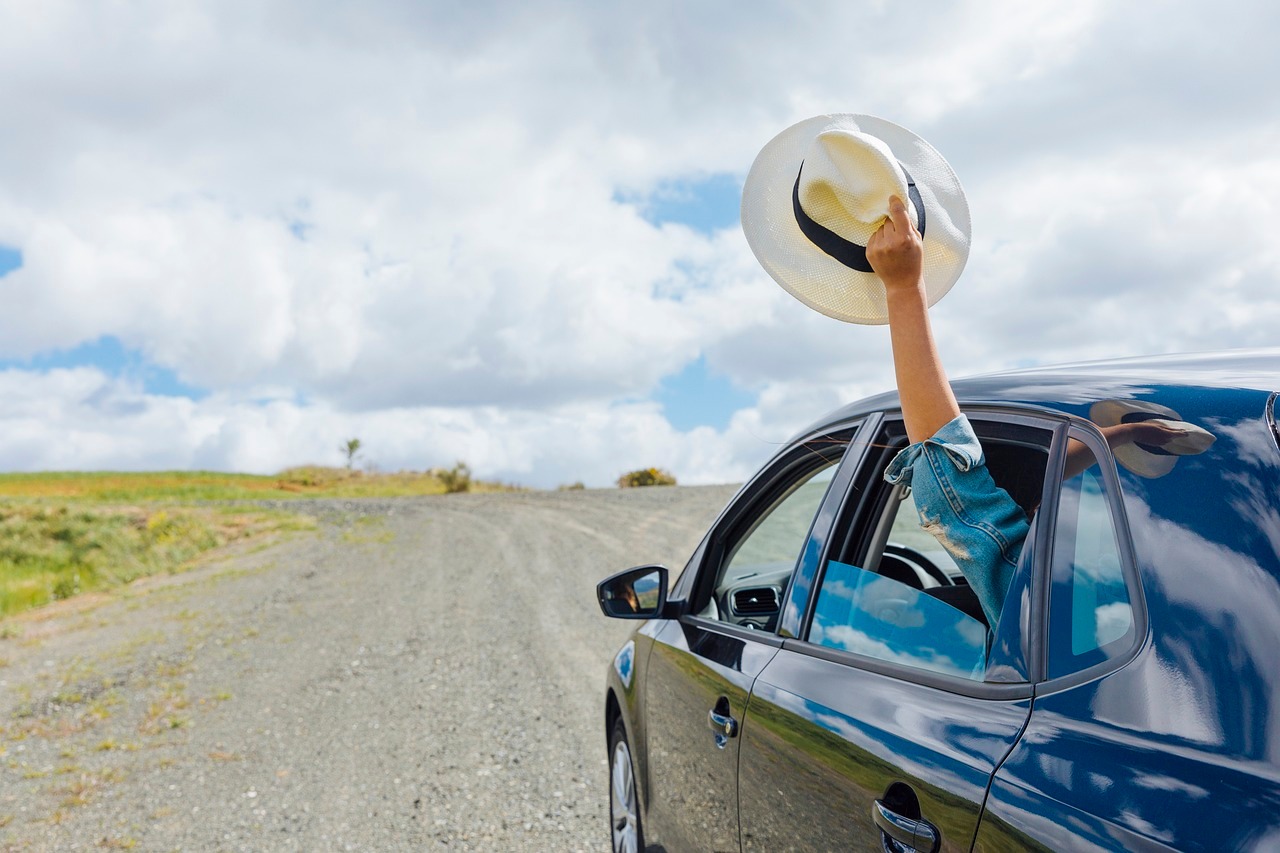 Ein Auto fährt in Richtung Horizont. Ein winkender Arm mit Hut in der Hand hängt aus dem Fenster