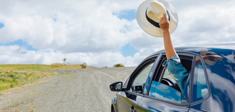 Ein Auto fährt in Richtung Horizont. Ein winkender Arm mit Hut in der Hand hängt aus dem Fenster