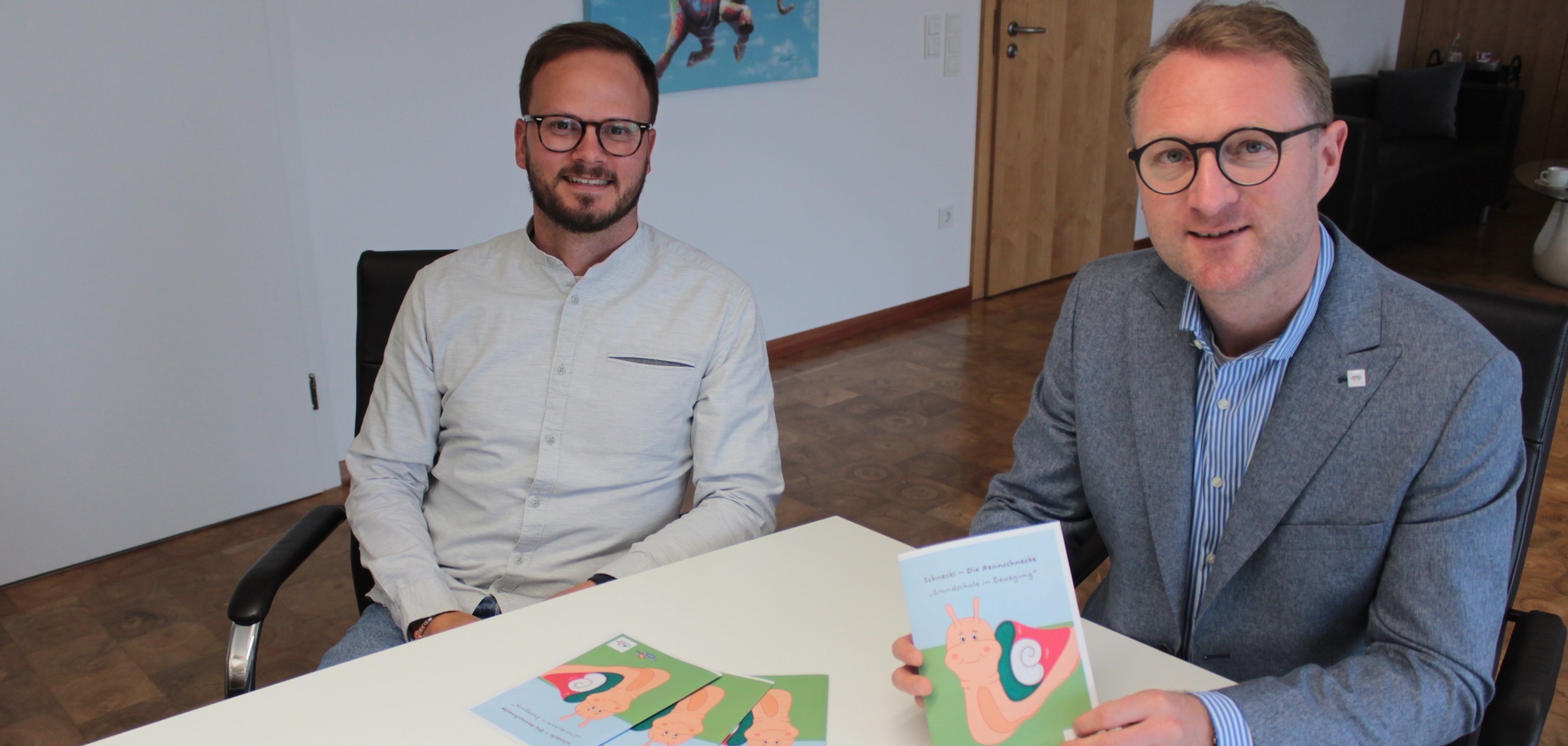 Zwei Männer halten einen Flyer in die Kamera und sitzen an einem weißen Konferenztisch. Sie machen auf ein Bewegungsprogramm für Grundschulkinder aufmerksam.