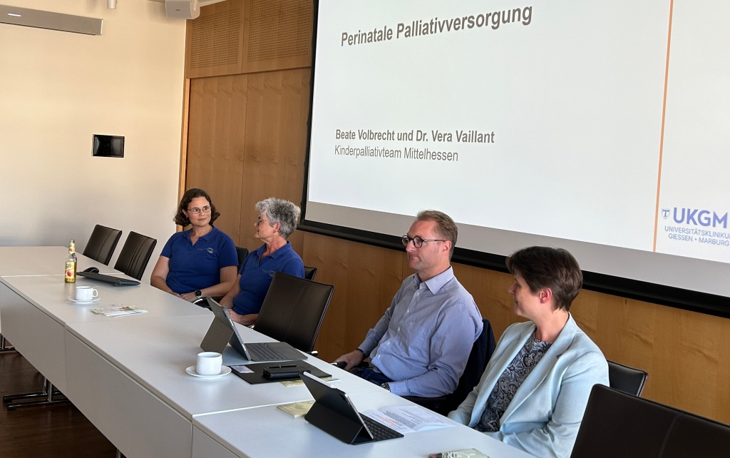Vier Personen sitzen an Tischen vor einer Leinwand. Im Hintergrund ist eine Präsentation zu sehen.